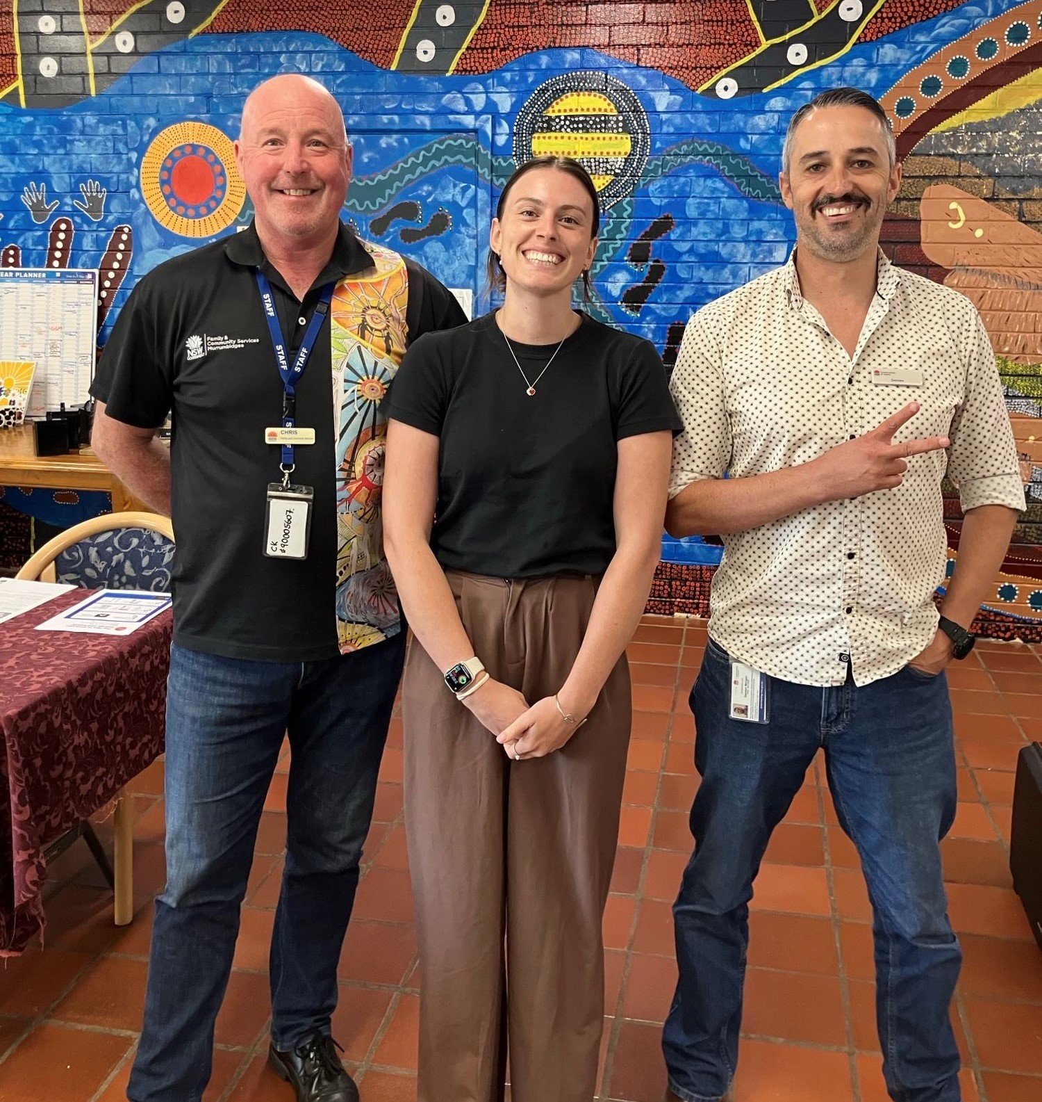 3 RMRA representatives smile in front of a mural featuring Aboriginal artwork
